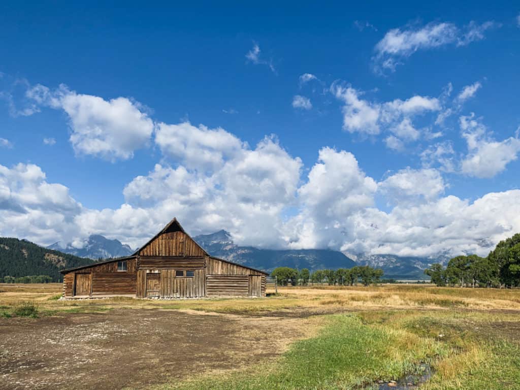 iconic wooden barn surrounded by grass and dirt against the backdrop of the majestic grand tetons under blue partly cloudy skies