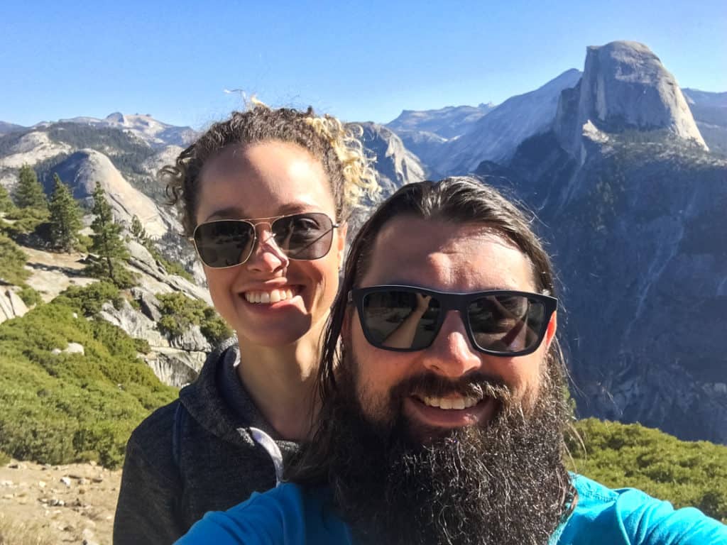 man with beard and brown hair and woman with brown curly hair in ponytail both wearing sunglasses and taking a selfie in front of Half Dome