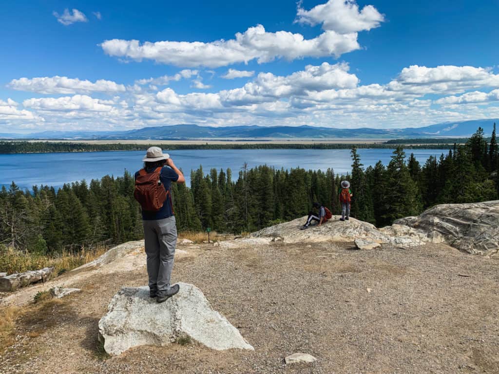 man standing on rock and looking through binoculars out across lake viewpoint while two boys play nearby