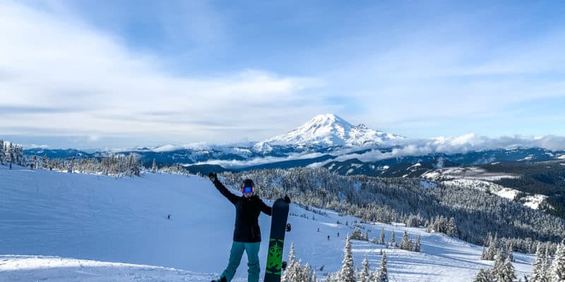 woman standing on snowy ridge in front of Mt. Rainier holding up a snowboard