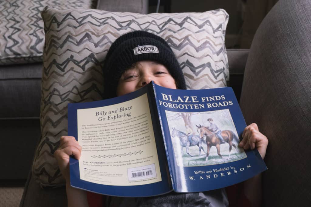 boy laying on couch reading book from Blaze series peeking out from behind book and smiling