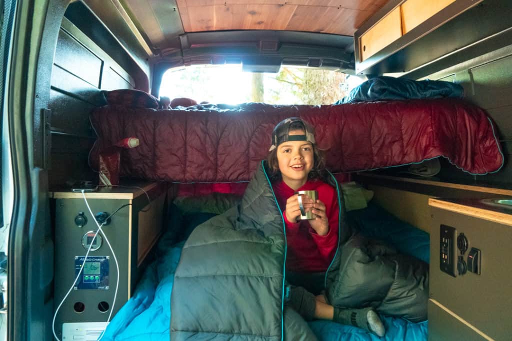 boy wrapped in blanket holding silver primus mug while camping in oregon at detroit lake in Roamerica campervan with Rumpl blankets on bed