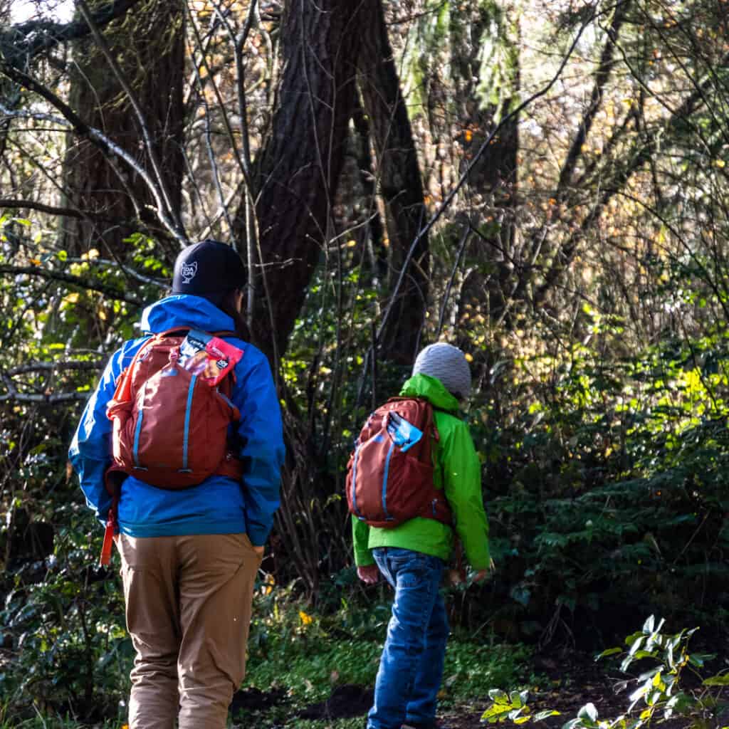 two boys hiking in the woods wearing brightly colored green and blue rain jackets while carrying rei backpacks on their backs