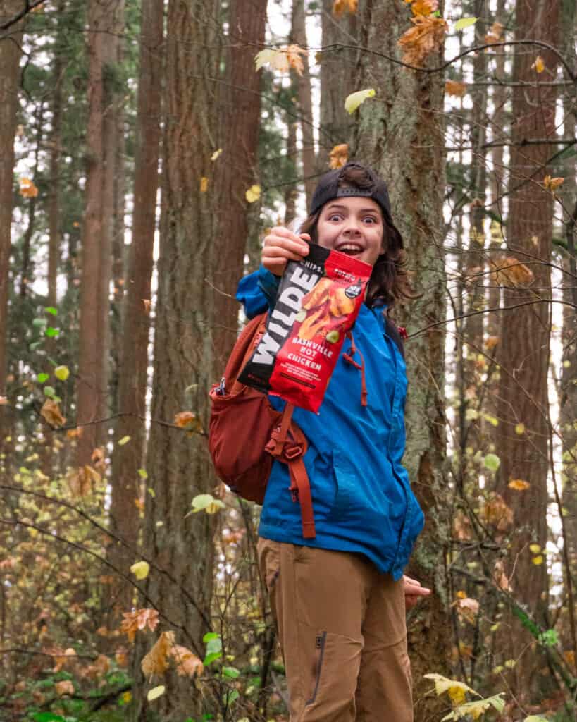 boy wearing blue rain jacket and tan hiking pants in among the trees and holding a bag of wilde chips in front of him with an excited look on his face