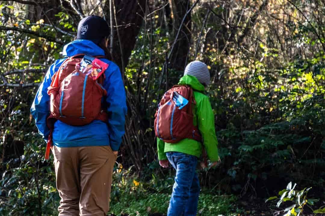 two boys hiking in the woods wearing brightly colored green and blue rain jackets while carrying rei backpacks on their backs