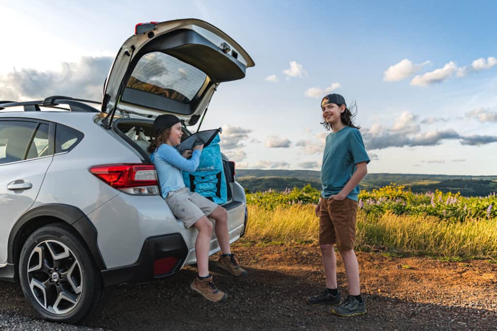 two boys smiling, one sitting under open hatch of white subaru reaches into icemule pro backpack cooler while the other is standing nearby