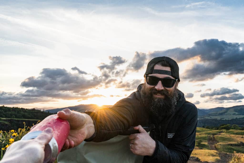 bearded man hands drink out of icemule boss backpack cooler to someone off camera with sunset in the background
