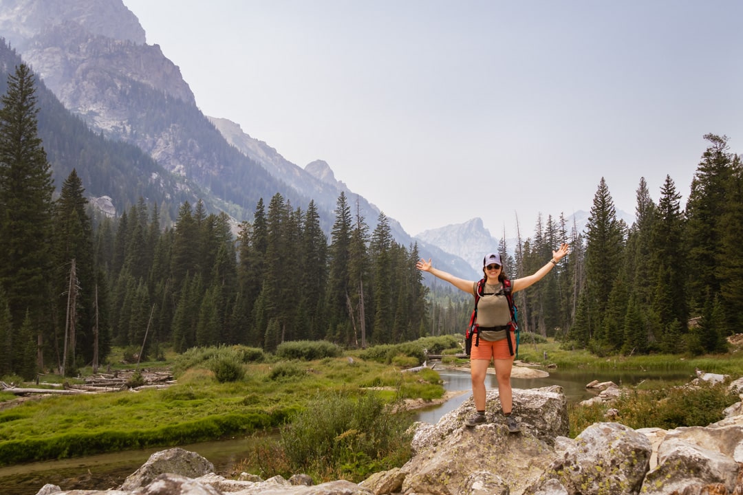 woman stands on rock next to river with Tetons in background