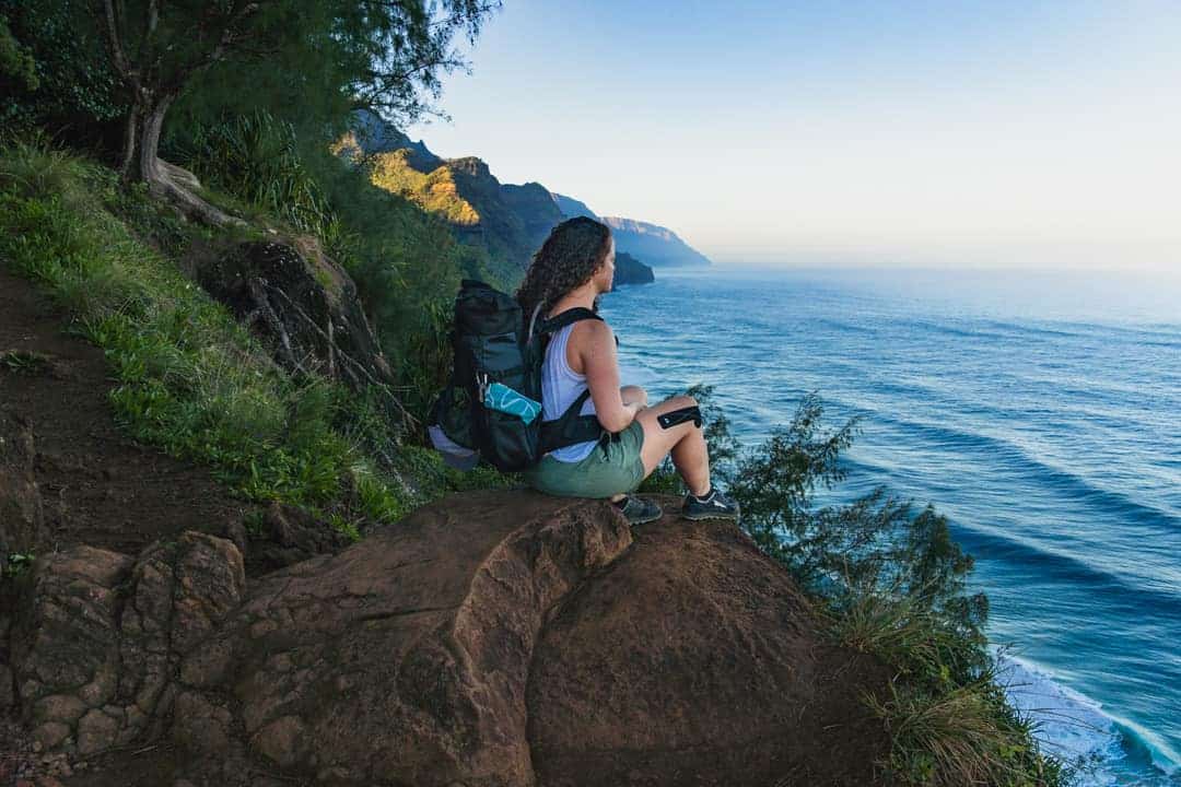 woman sits looking out over ocean along kalalau trail in kauai
