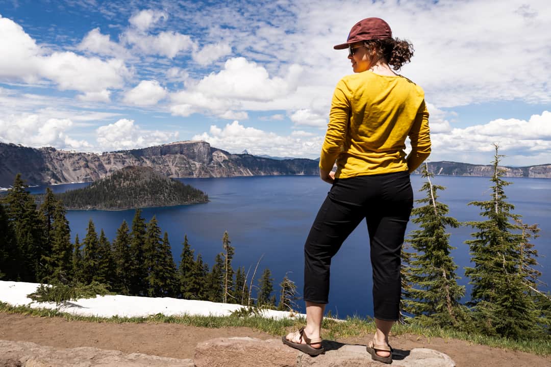 woman stands in on edge of trail at crater lake national park wearing black hiking pants, yellow hiking shirt, and hat