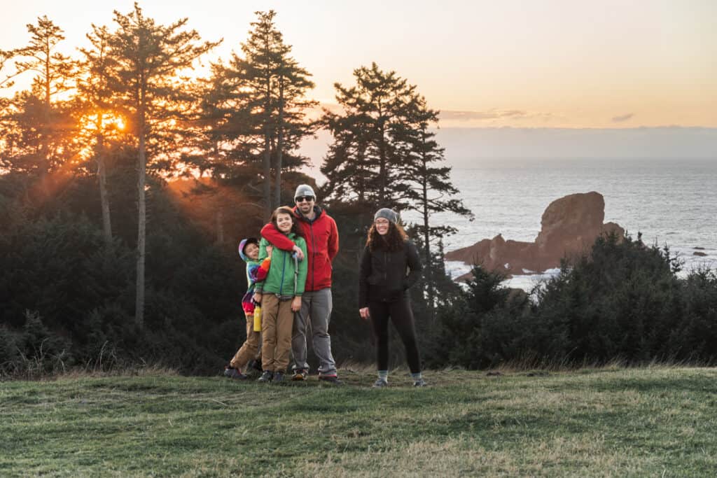 family stands smiling and laughing in front of coastline in Oregon wearing rain jackets at sunset