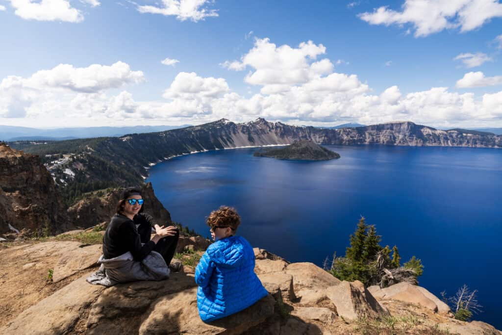 two boys wearing sunglasses sit at edge of trail along hike at crater lake