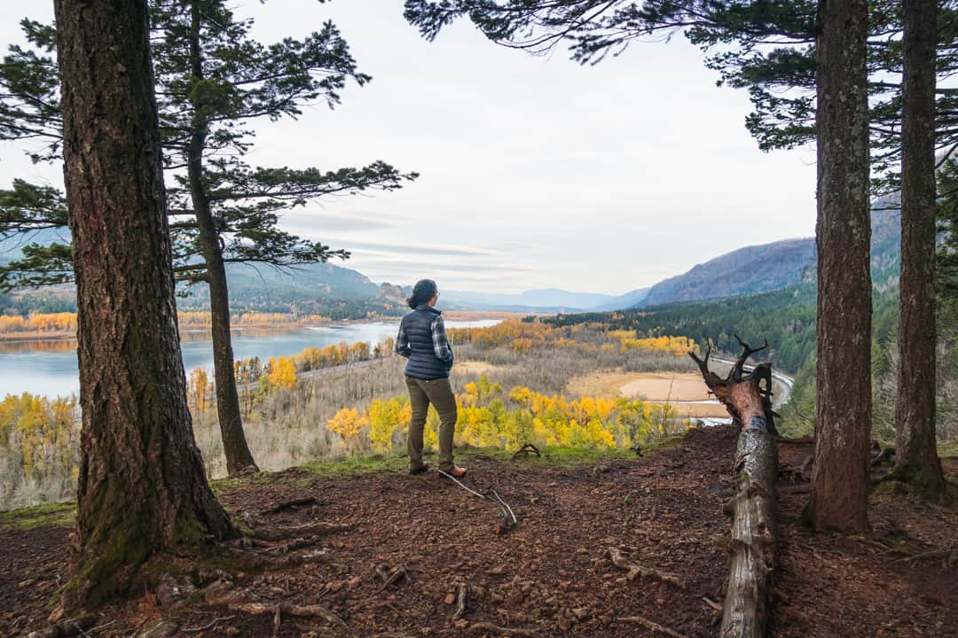 woman stands looking out at columbia river gorge on a hike wearing a puffy vest, hiking pants, and hiking boots
