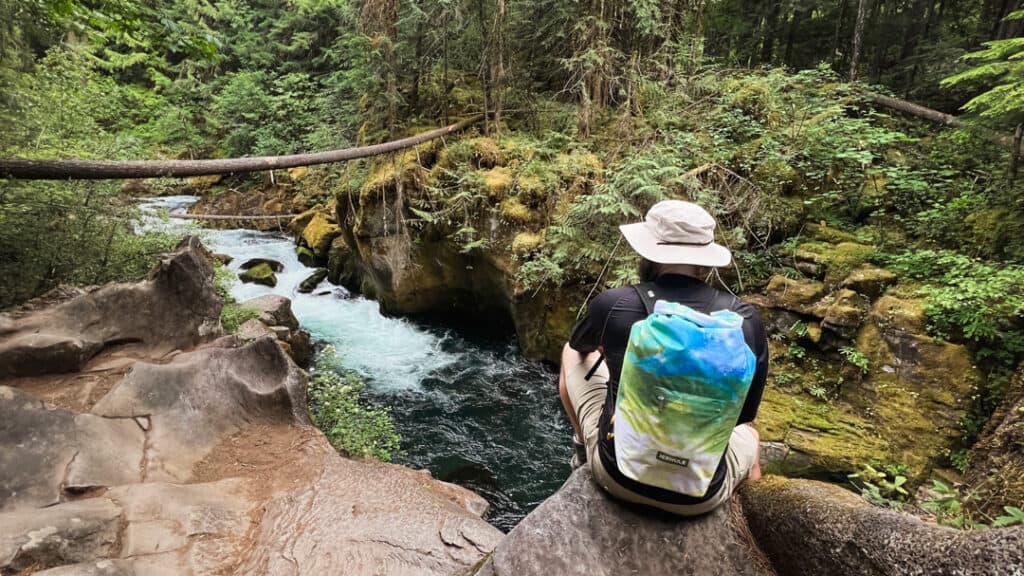 man sits next to river in beige hat wearing icemule backpack cooler during a hike