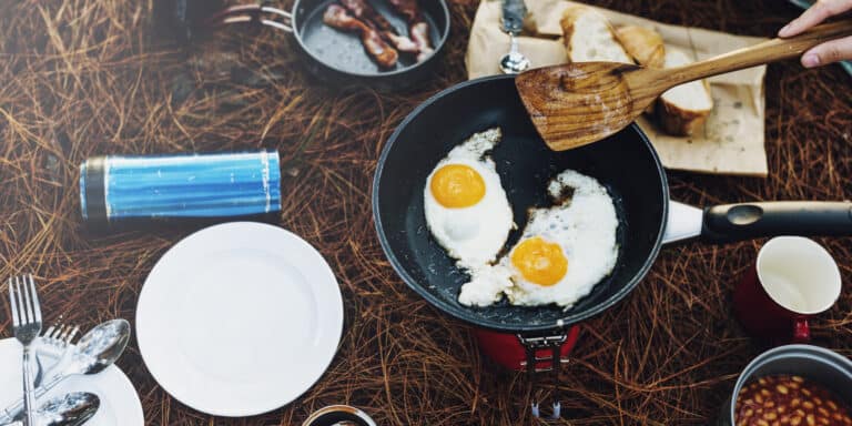 frying pan sits on top of small backpacking stove with two fried eggs and a hand holding a wooden spoon with other camping dishes and food surrounding the stove