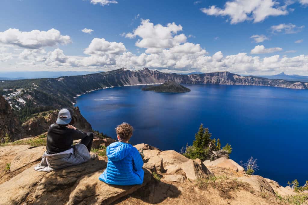 two boys sit on top of a rock looking over Crater Lake, one of the National Parks in Oregon and Washington