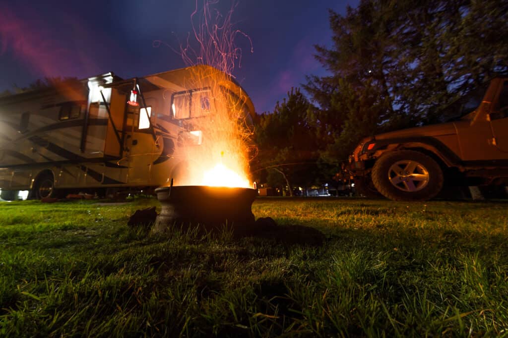 a fire burns brightly at an RV campsite with rv and jeep in the background