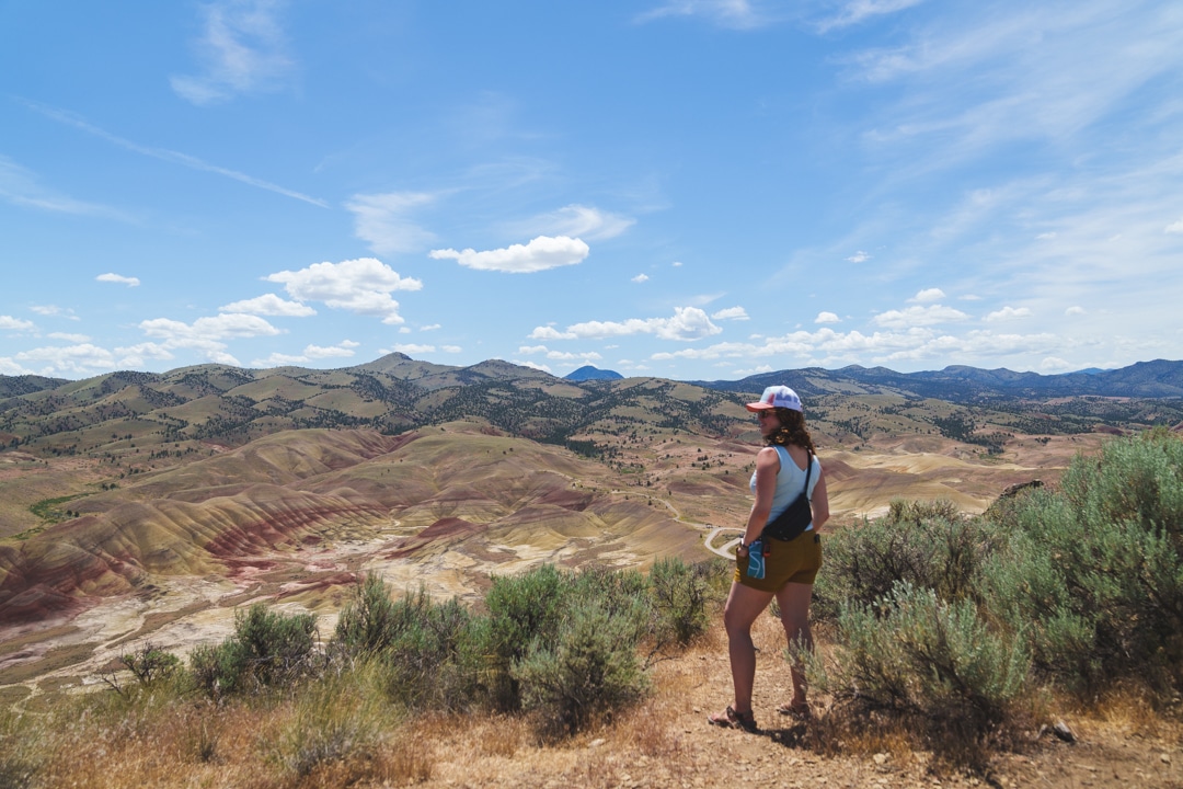 A woman stands at a vantage point at the top of the Carroll Rim Trail in the Painted Hills Oregon. She is wearing shorts, hiking sandals, a light colored tank top, a hat, and sunglasses for sun protection under the sunny, partly cloudy skies and is carrying additional essentials in a fanny pack.