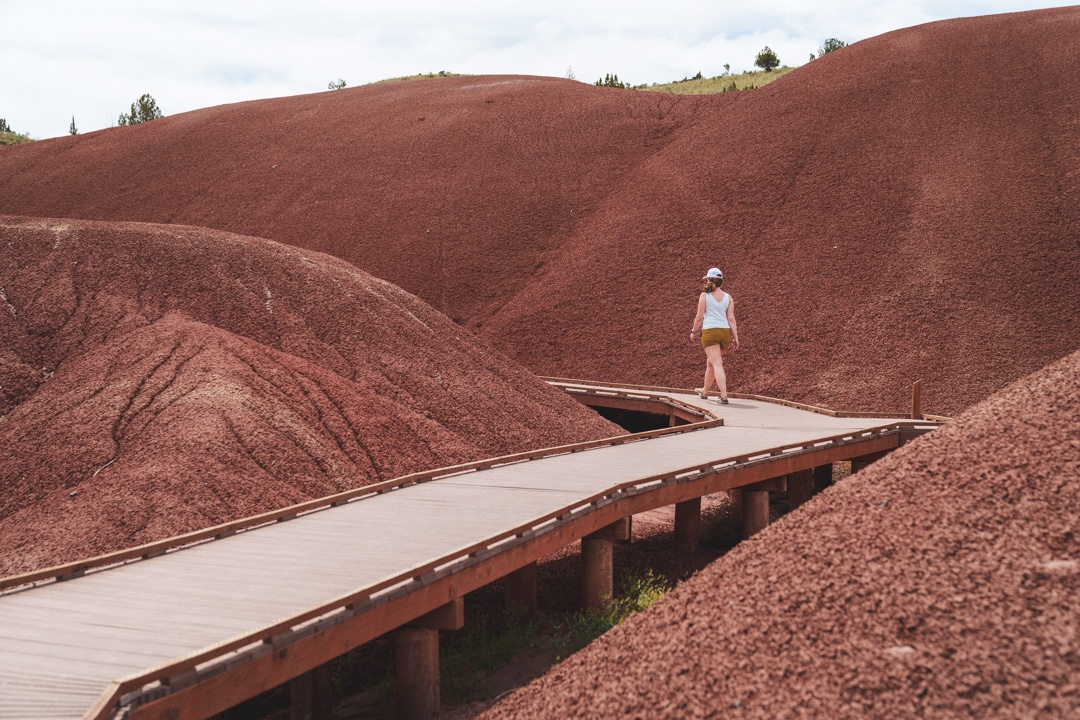 A woman walks on a boardwalk through the bright red colors of the Painted Hills on the Painted Cove Trail. She is wearing shorts, hiking sandals, a light colored tank top, a hat, and sunglasses for sun protection under the sunny, partly cloudy skies. 