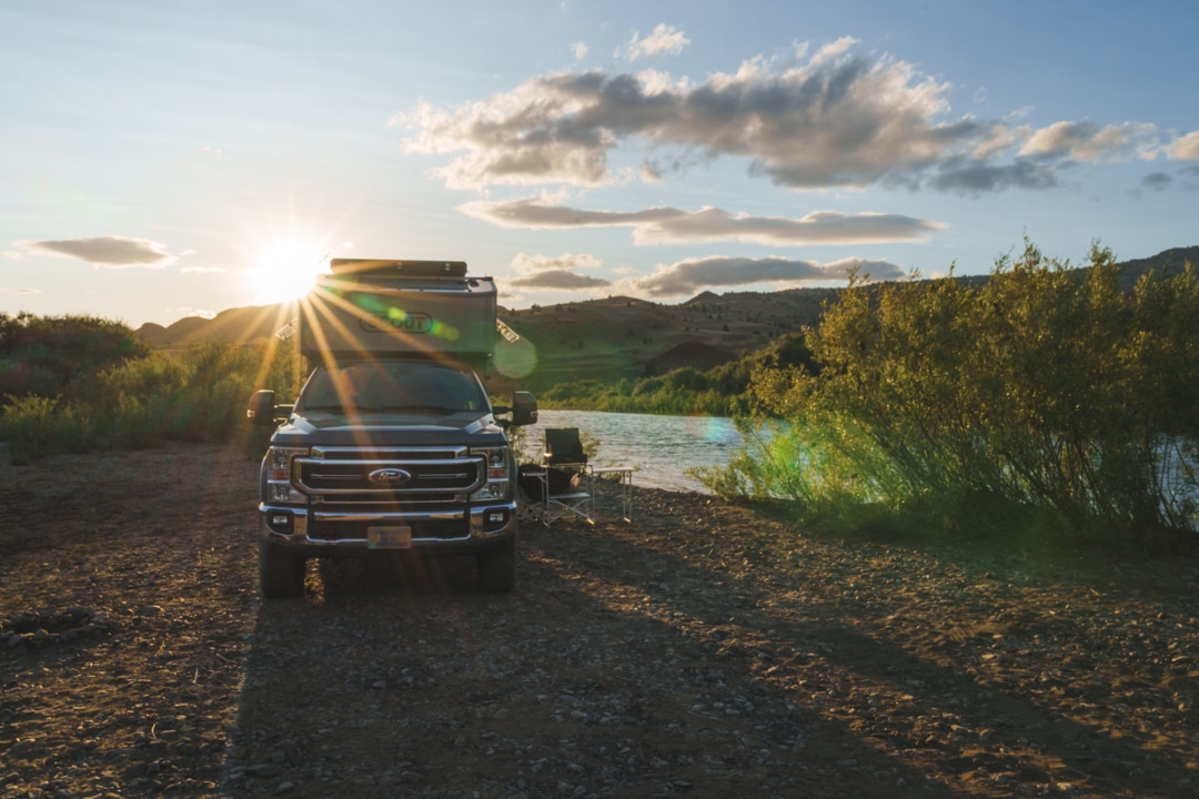 A Ford F-350 with a Scout Kenai camper sits at a campsite at Priest Hole Recreation Site near John Day National Monument. There is a river running alongside the campsite and a sunburst is glowing at the crest of the hills behind the camper during golden hour.