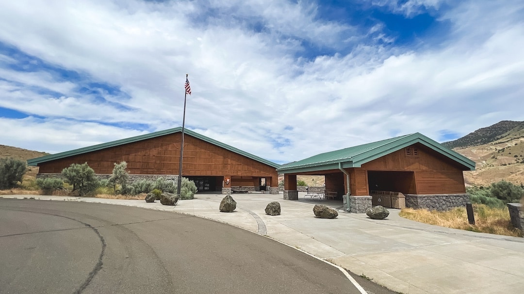 The Thomas Condon Visitor Center sits under a blue cloudy sky. The building is in tones of green and brown and there is a flag pole with the American Flag flying out front. A driveway curves around in front of the building.