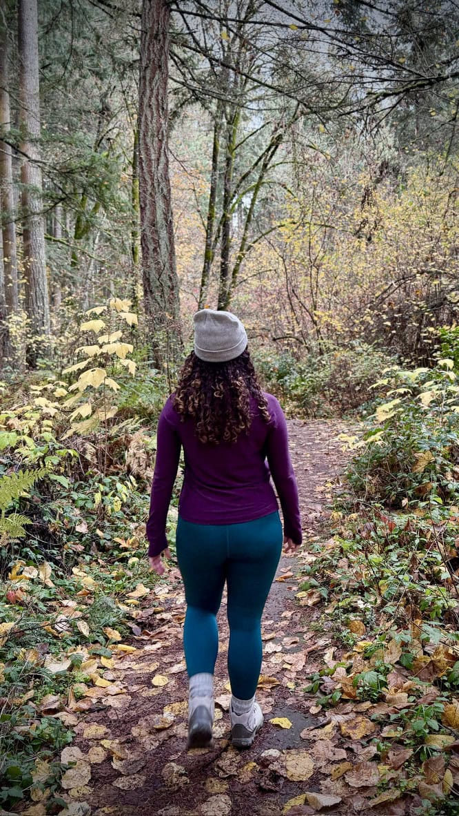 A woman walking away on a dirt path surrounded by fall foliage, dressed in a mulberry heather Title Nine Grace Raglan Long Sleeve Tee and dark sea Kirsi Tight. She completes the outfit with a cozy gray beanie and hiking boots.