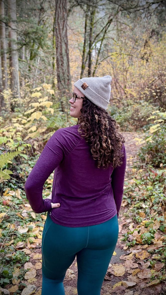 Back view of a woman hiking on a leafy forest trail in fall, wearing a mulberry heather Grace Raglan Long Sleeve Tee, dark sea Kirsi Tight, and a gray beanie. The colorful autumn foliage adds a scenic backdrop.