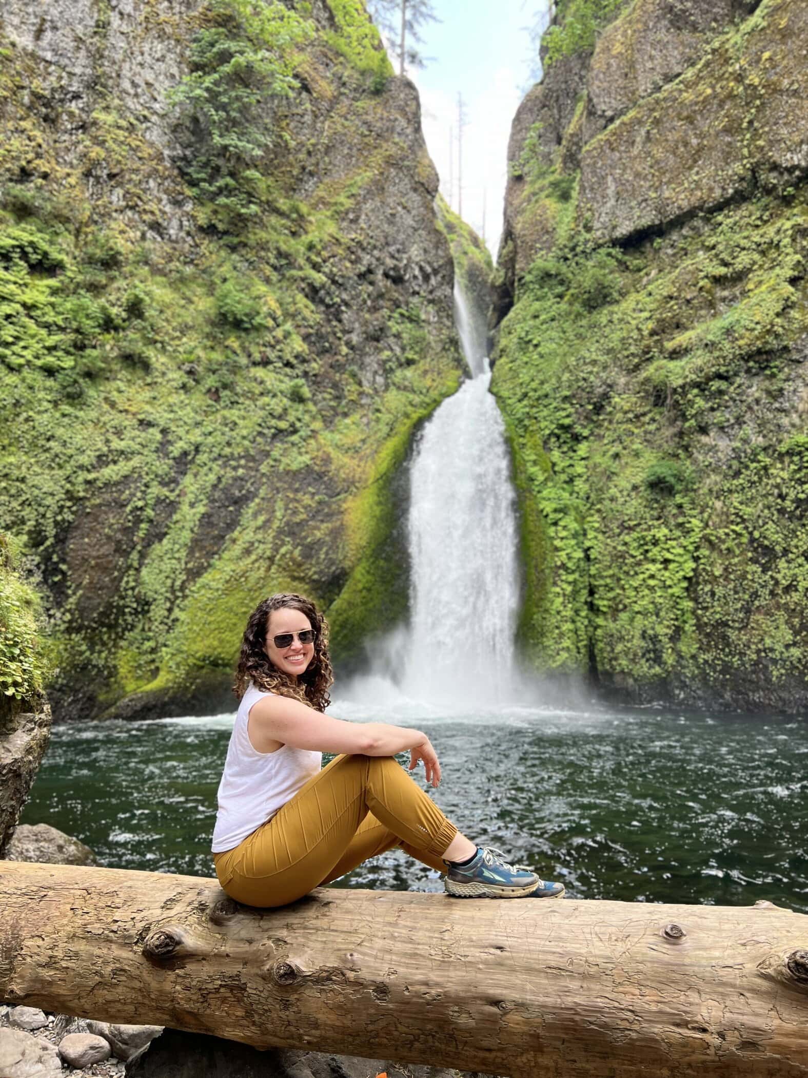 A smiling hiker sits on a log with Wahclella Falls flowing behind her into a serene pool, surrounded by lush mossy cliffs. This popular viewpoint near the end of the trail offers a scenic and photogenic backdrop.