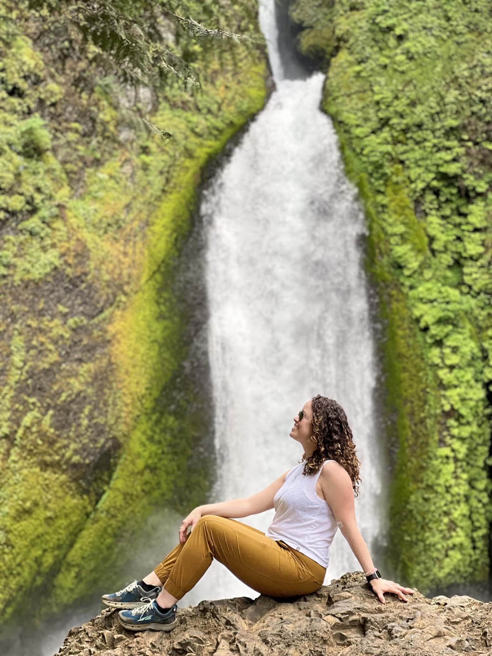 Wahclella Falls Trail: Easy & Stunning Columbia Gorge Hike 8 A woman poses on a rocky ledge in front of Wahclella Falls, with the powerful cascade framed by vibrant green moss on the canyon walls. The dramatic cliffside angle makes this a striking photo spot along the trail.